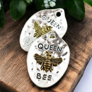 May include: Two silver-colored, teardrop-shaped pendants with a hammered texture. Each pendant features a bronze-colored bee and the word "QUEEN" above and "BEE" below. The pendants are on a wooden surface, with greenery in the background.
