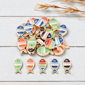 May include: A collection of colorful fish-shaped charms with gold-colored edges. The charms are painted with stripes in various colors, including red, blue, green, and pink. The charms are arranged on a white wooden surface.