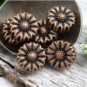 May include: A group of seven copper-colored flower-shaped beads with black accents. The beads are arranged in a bowl on a wooden surface.