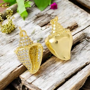 May include: Two gold-colored seashell pendants. One pendant is encrusted with clear crystals, while the other is a solid, polished gold. Both have a loop for hanging. The pendants rest on weathered wood.