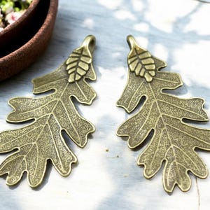 May include: Two bronze-colored oak leaf pendants with detailed veining and a small acorn at the top. The pendants are designed with a loop for hanging and are ideal for jewelry making or crafting projects.