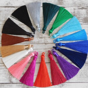 May include: A collection of 15 colorful silk tassels, each with a different color, arranged in a circle on a white wooden surface. The tassels are made of soft, silky material and have a fringe at the bottom.