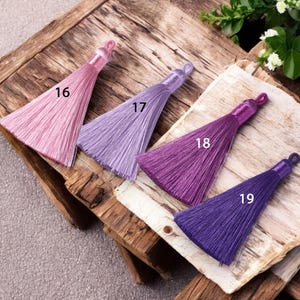 May include: Four silk tassels in shades of pink, lavender, purple, and deep purple. The tassels are arranged in a row on a wooden surface, with the numbers 16, 17, 18, and 19 printed next to each tassel.