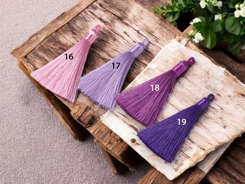 May include: Four silk tassels in shades of pink, lavender, purple, and deep purple. The tassels are arranged in a row on a wooden surface, with the numbers 16, 17, 18, and 19 printed next to each tassel.