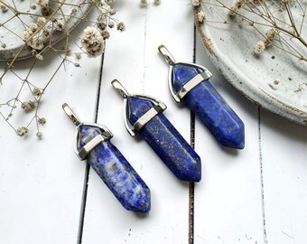 May include: Three blue lapis lazuli pendants with silver-colored metal caps. The polished, hexagonal pendants are arranged on a white wooden surface, with dried flowers and a ceramic plate in the background. The pendants are approximately 2 inches long.