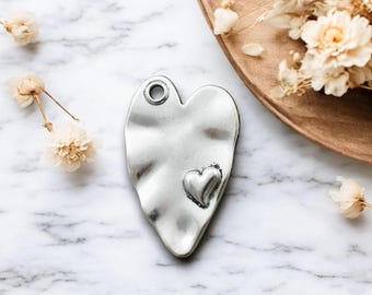May include: A silver-colored heart-shaped pendant with a smaller heart detail. The pendant has a textured, hammered appearance and a hole at the top for hanging. The background includes dried flowers and a wooden tray.