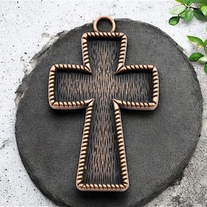 May include: A large, ornate, copper-colored cross pendant with a black, textured center. The cross has a rope-like border and a loop at the top for hanging.
