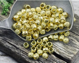 May include: A large pile of gold-colored metal beads with a flower-like design. The beads are in a metal spoon on a wooden surface.