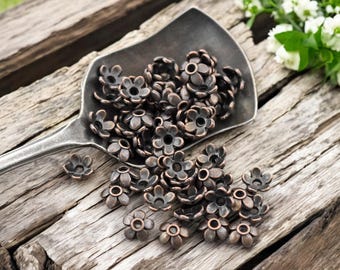 May include: A pile of antique copper flower-shaped bead caps, scattered on a rustic wooden surface. The bead caps are ideal for adding a touch of vintage charm to jewellery making projects.