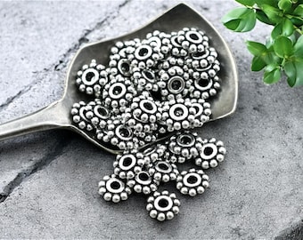 May include: A pile of silver metal beads with a raised circle design. The beads are arranged on a silver spoon.