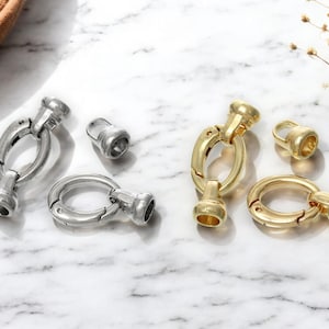 May include: Close-up of jewelry clasps in silver and gold tones. The clasps are oval-shaped with a hinged design, and there are also smaller, cylindrical components. The items are displayed on a marble surface, suggesting a focus on jewelry making or accessories.