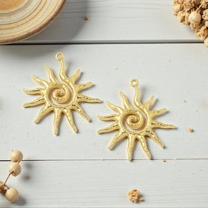 May include: Two gold-colored sun-shaped pendants with a spiral center and pointed rays. The pendants are on a white wooden surface, with a small dish and dried flowers in the background. These pendants are suitable for jewelry making.