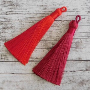 May include: Two tassels, one red and one burgundy, are shown against a white wood background. The tassels are made of soft, silky threads and have a loop at the top for attaching.