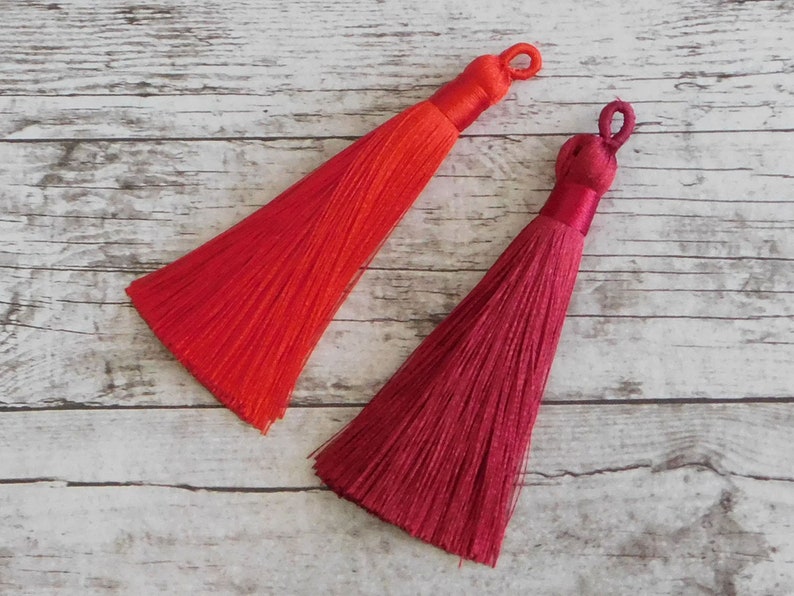 May include: Two tassels, one red and one burgundy, are shown against a white wood background. The tassels are made of soft, silky threads and have a loop at the top for attaching.