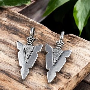 May include: Two silver arrowhead pendants with a textured surface and a geometric design. The pendants are on a wooden surface.