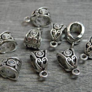 May include: Eight silver-toned metal pendant caps with ornate designs. The caps have a loop at the top for attaching to a chain or cord.