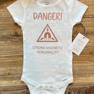 May include: White baby onesie with a rose gold graphic that says "DANGER!" with a triangle containing a magnet and lightning bolts. The text below the triangle says "STRONG MAGNETIC PERSONALITY".