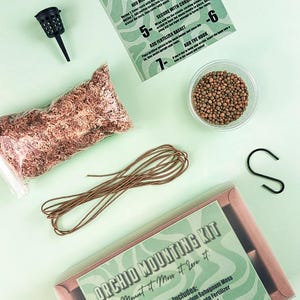 Op de afbeelding: Een orchideeën montagekit met instructies, veenmos, meststof, een mandje, koord en een S-haak. De kit bevat een zwarte plastic mand, een doorzichtige bak met meststof en een verpakking mos. De kit is ontworpen om orchideeën te monteren.