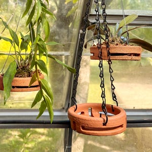 May include: Three circular terracotta hanging planters with horizontal side slits, suspended by black metal chains. Two planters contain green leafy plants with visible roots and soil, while one empty planter hangs in the foreground.