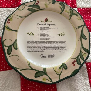 May include: A white ceramic plate with a green floral design and a handwritten recipe for Caramel Popcorn. The recipe includes ingredients like popped popcorn, peanuts, pumpkin seeds, butter, brown sugar, corn syrup, and maple syrup. The recipe is signed "Gaul Allen".