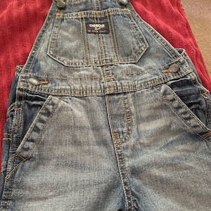 May include: Blue denim overalls with adjustable straps and a brown leather patch with the text "OSHKOSH B'Gosh" and "The Original Overall".