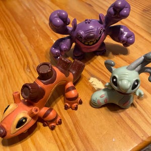 May include: Three character figurines in vibrant colours. A purple figure with large arms and a wide grin, an orange and brown figure with a unique shape, and a light green figure with large ears and pink accents.