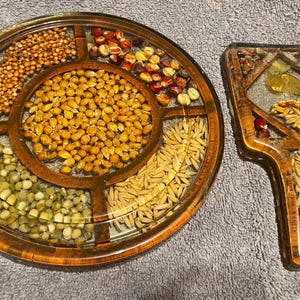 May include: A round, wooden-framed serving tray with six compartments, each filled with different grains and seeds. Next to it is a matching serving utensil, also with grains and seeds embedded in resin. The tray measures approximately 12 inches in diameter.