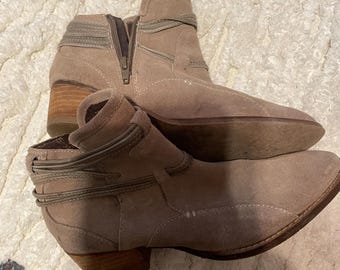 May include: A pair of taupe suede ankle boots with a stacked wooden heel. The boots feature a side zipper and decorative straps around the ankle. The heel height is approximately 2 inches.