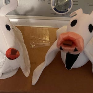 May include: Two plush toys of Zero from The Nightmare Before Christmas. The toys are white with black eyes, a red nose, and a pink mouth. One has a sheer white wing.