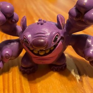 May include: A purple Stitch figurine with large ears, a wide grin, and raised arms. The figure has a pink mouth and yellow teeth. The figurine is on a wooden surface.