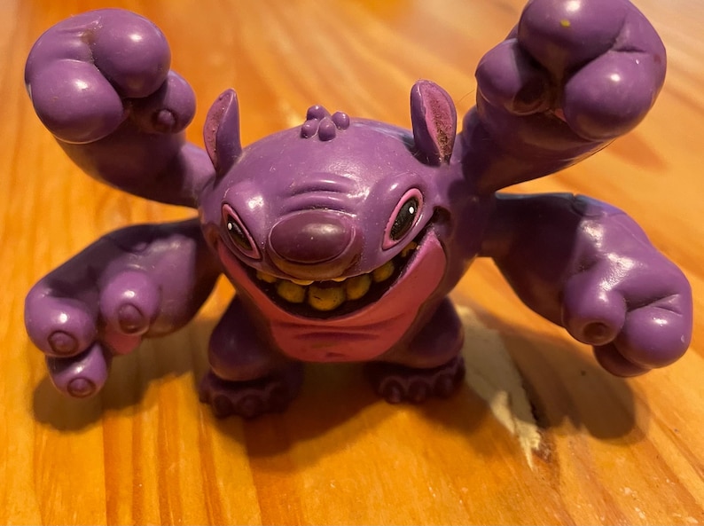 May include: A purple Stitch figurine with large ears, a wide grin, and raised arms. The figure has a pink mouth and yellow teeth. The figurine is on a wooden surface.