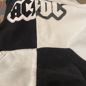 May include: Black and white hoodie with the band name "AC/DC" in a white, outlined font. The hoodie is split down the middle, with one side black and the other white. The design is a bold statement.