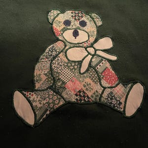 May include: Dark green sweatshirt featuring a patchwork teddy bear design. The bear is made of various patterned fabrics in shades of green, red, and cream, with a white bow and accents. The bear is sitting with its legs spread.