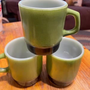 May include: Three green glass mugs with a white interior and a dark brown rim. The mugs are stacked on top of each other.