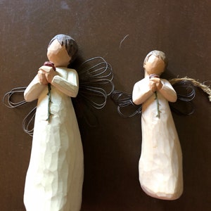 May include: Two white wooden angel figurines holding a single red rose. The angels have wire wings and are standing on a brown surface.