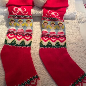 May include: Two red Christmas stockings with white pom-poms, featuring a knitted design of angels, bows, and geometric patterns in white, green, and yellow. The stockings have a white cuff and toe.