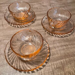 May include: Three pink glass teacups and saucers with a swirl design. The cups and saucers are all the same size and shape.