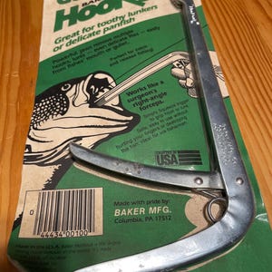 May include: A silver Hookout fishing tool on a green and white package. The package reads "Get The Hookout" and describes the tool's use for removing hooks from fish. Made in the USA.