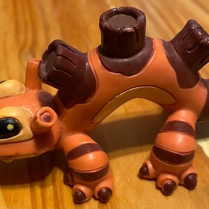 May include: A plastic figurine in orange and brown, depicting a cartoon creature. It has large eyes, a unique head shape, and dark brown patches and stripes. The figure features a cylindrical shape on its back.