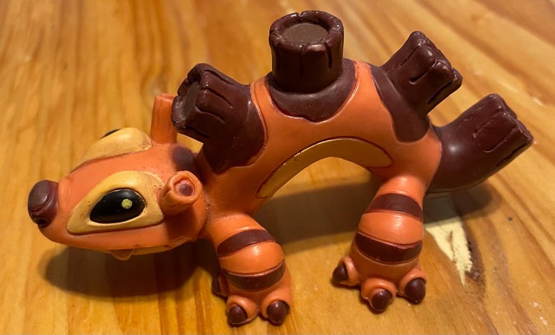 May include: A plastic figurine in orange and brown, depicting a cartoon creature. It has large eyes, a unique head shape, and dark brown patches and stripes. The figure features a cylindrical shape on its back.