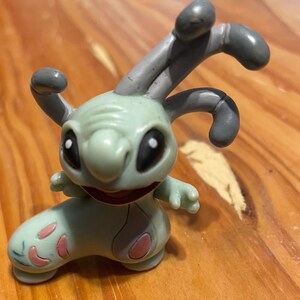 May include: A light green cartoon figure with large black eyes, a wide smile, and grey antennae. The figure has pink accents on its feet and is posed on a wooden surface. The toy is a collectible item.