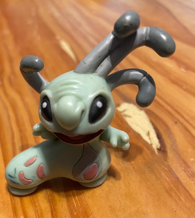 May include: A light green cartoon figure with large black eyes, a wide smile, and grey antennae. The figure has pink accents on its feet and is posed on a wooden surface. The toy is a collectible item.