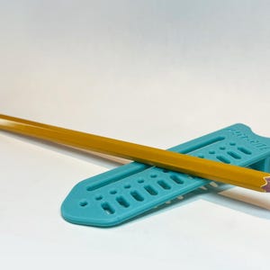 May include: A yellow pencil with a pink eraser rests on a teal-colored pencil holder. The pencil holder has a series of rectangular cutouts and the text "Paper-Mate" printed on it. The pencil is sharpened and ready for use.