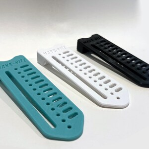 May include: Three plastic MARK-RITE tools in teal, white, and black. Each tool has a rectangular shape with a series of holes and the brand name printed on the top. The tools are designed for marking or measuring.