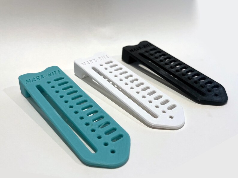 May include: Three plastic MARK-RITE tools in teal, white, and black. Each tool has a rectangular shape with a series of holes and the brand name printed on the top. The tools are designed for marking or measuring.