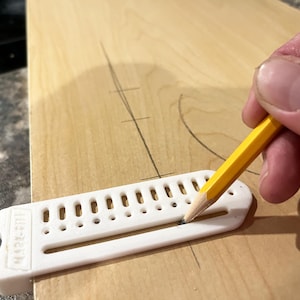 May include: A yellow pencil is used to mark a wooden surface with a white plastic marking tool. The tool has a series of holes and the text "MARK-RITE" is visible. The wood has pencil markings.