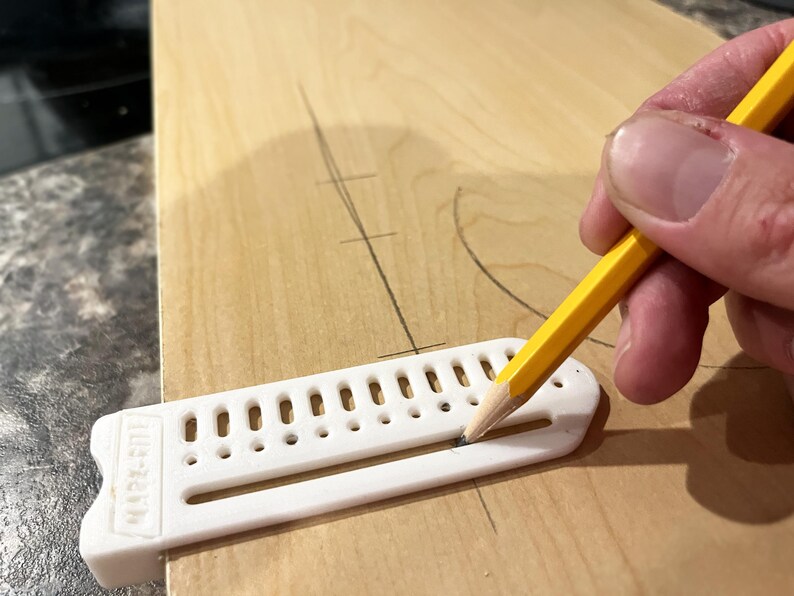 May include: A yellow pencil is used to mark a wooden surface with a white plastic marking tool. The tool has a series of holes and the text "MARK-RITE" is visible. The wood has pencil markings.