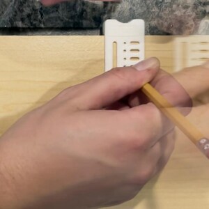 May include: A hand using a yellow pencil to draw on a light-colored wooden surface. A white plastic guide is attached to the wood. The pencil has a pink eraser.