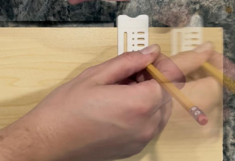 May include: A hand using a yellow pencil to draw on a light-colored wooden surface. A white plastic guide is attached to the wood. The pencil has a pink eraser.