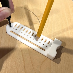 May include: A white plastic marking guide with a series of holes and slots is used with a pencil and a black marking tool on a wooden surface. The pencil is yellow with a gray tip.